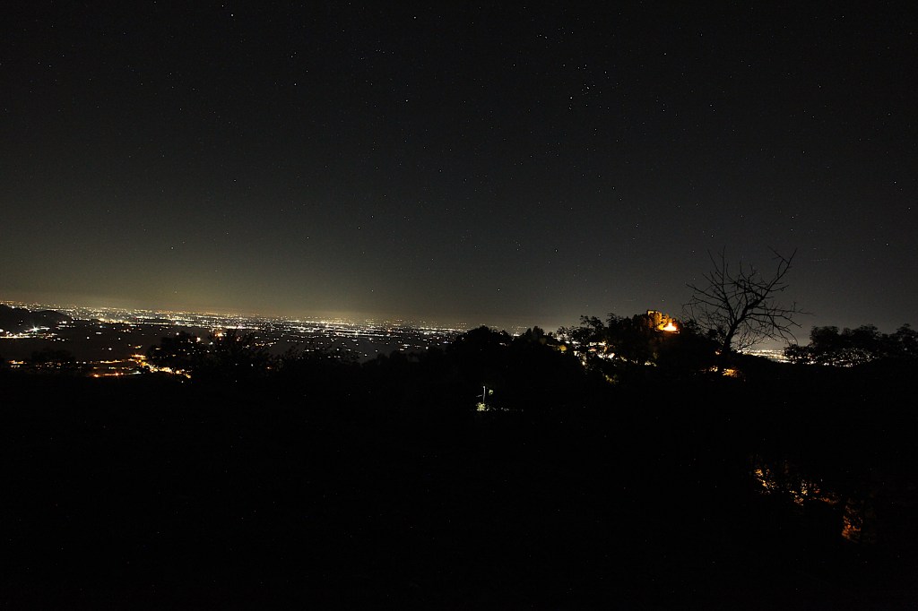 Panorama notturno di una città illuminata con una leggera aura di luci, visibile sullo sfondo, con un castello in alto a destra e stelle nel cielo.